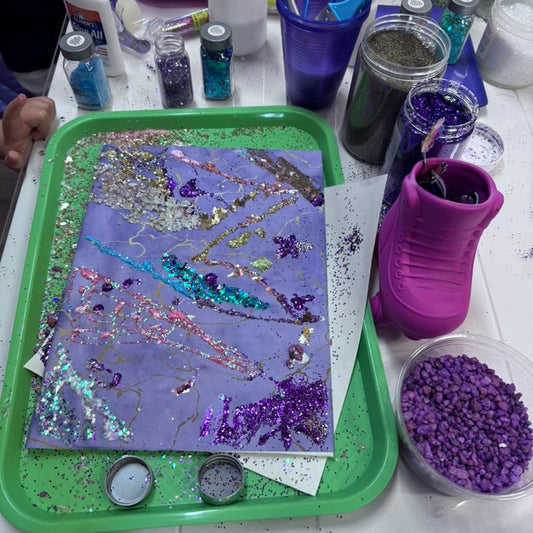 Artistic creation with glitter on a purple surface, surrounded by art supplies on a table.