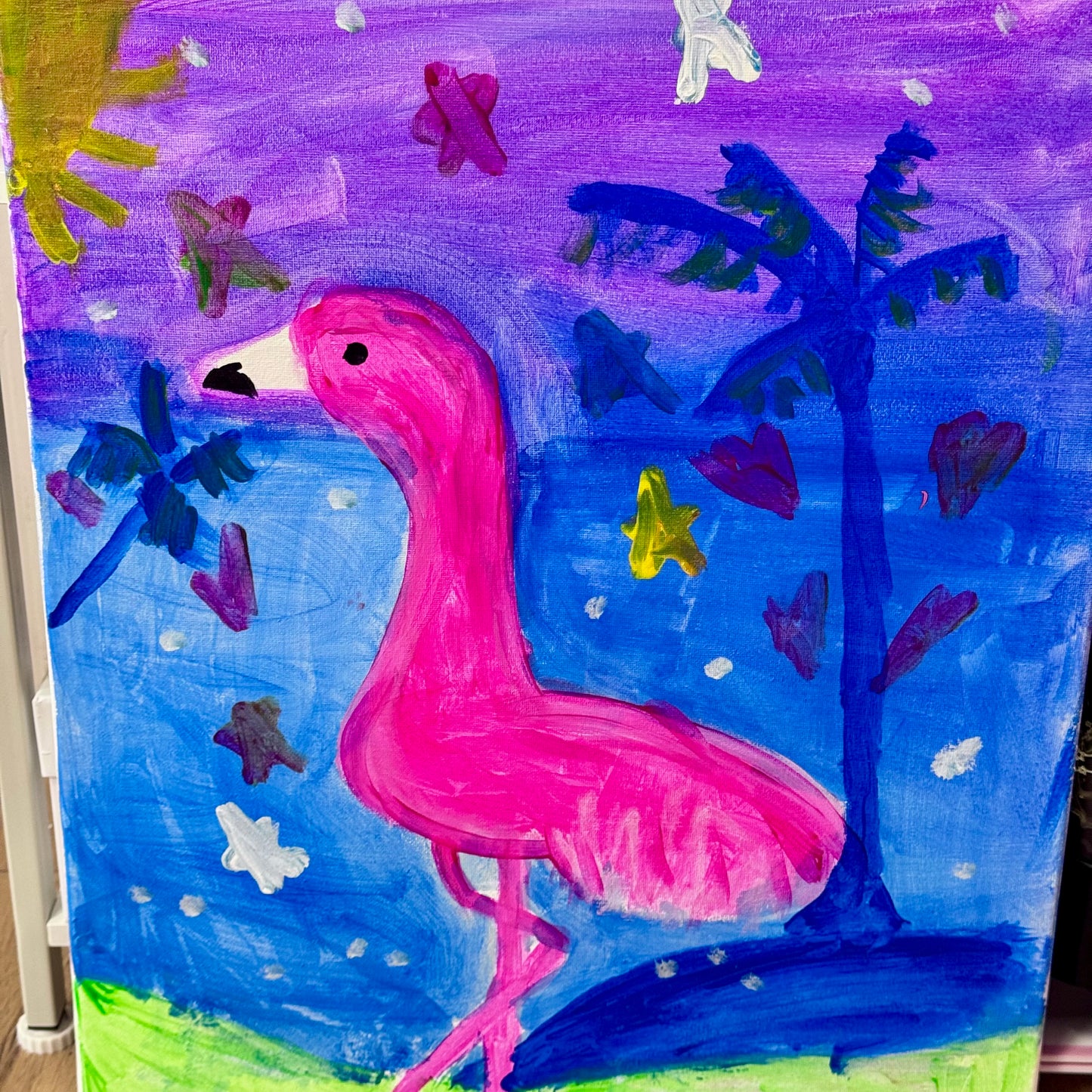 A colorful painted artwork featuring a bright pink flamingo standing on a blue and purple landscape with dark blue trees, green grass, and scattered white and yellow paint marks.