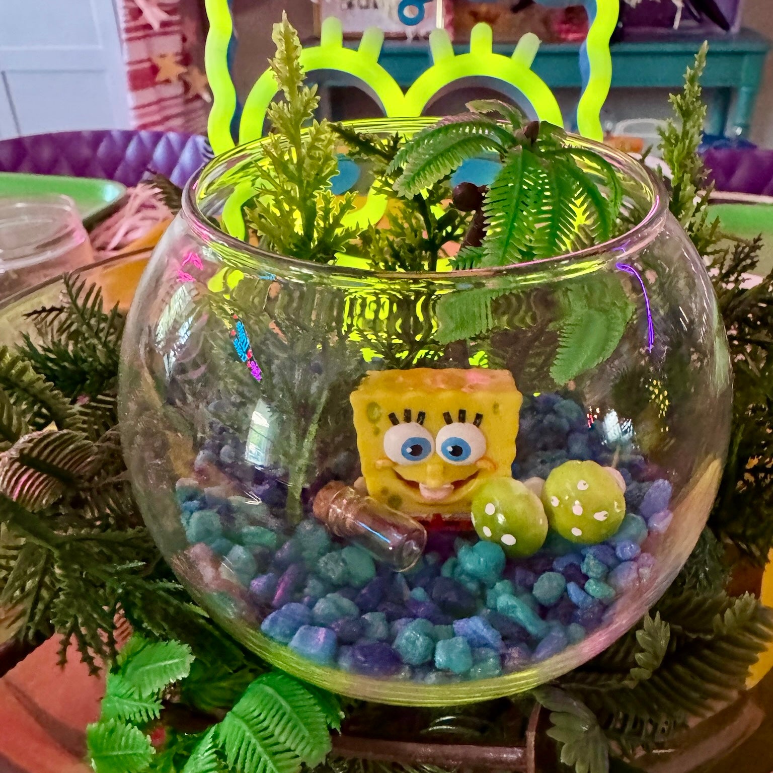 A clear glass fishbowl terrarium filled with blue and purple pebbles, green artificial ferns, and a SpongeBob character figure, set on a table with a neon green sign in the background.