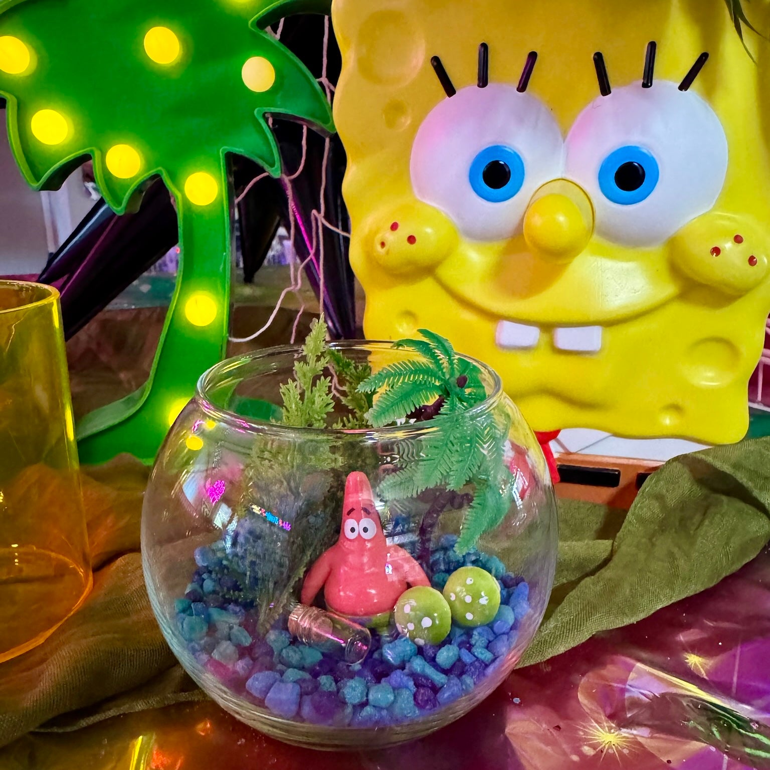 A clear glass fishbowl terrarium filled with blue and purple pebbles, a small pink starfish figure, green plants, and two small green eggs, styled in front of a large yellow SpongeBob face light and a green palm tree light.