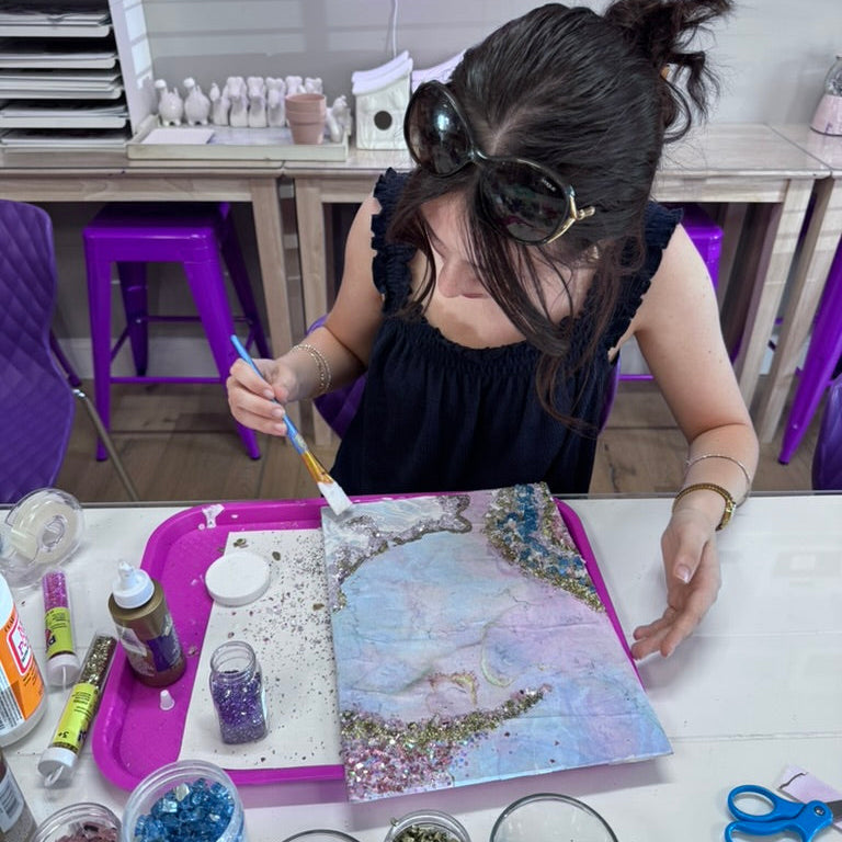 Person working on a craft project with glitter and paint supplies at a table.