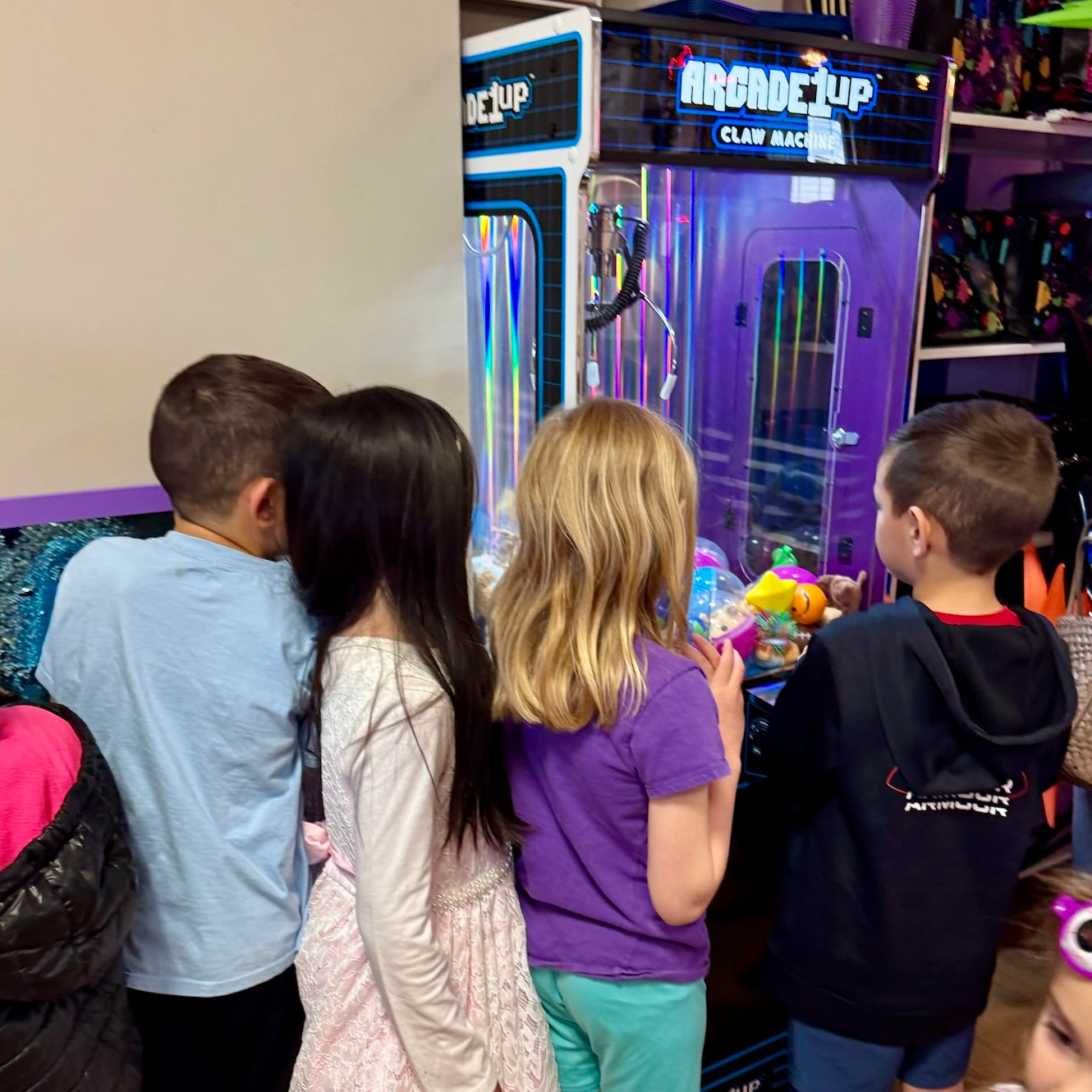 Four children standing in front of a claw machine in an indoor arcade or play area. The claw machine is purple with blue neon-style signage reading 'Arcade Cup Claw Machine.