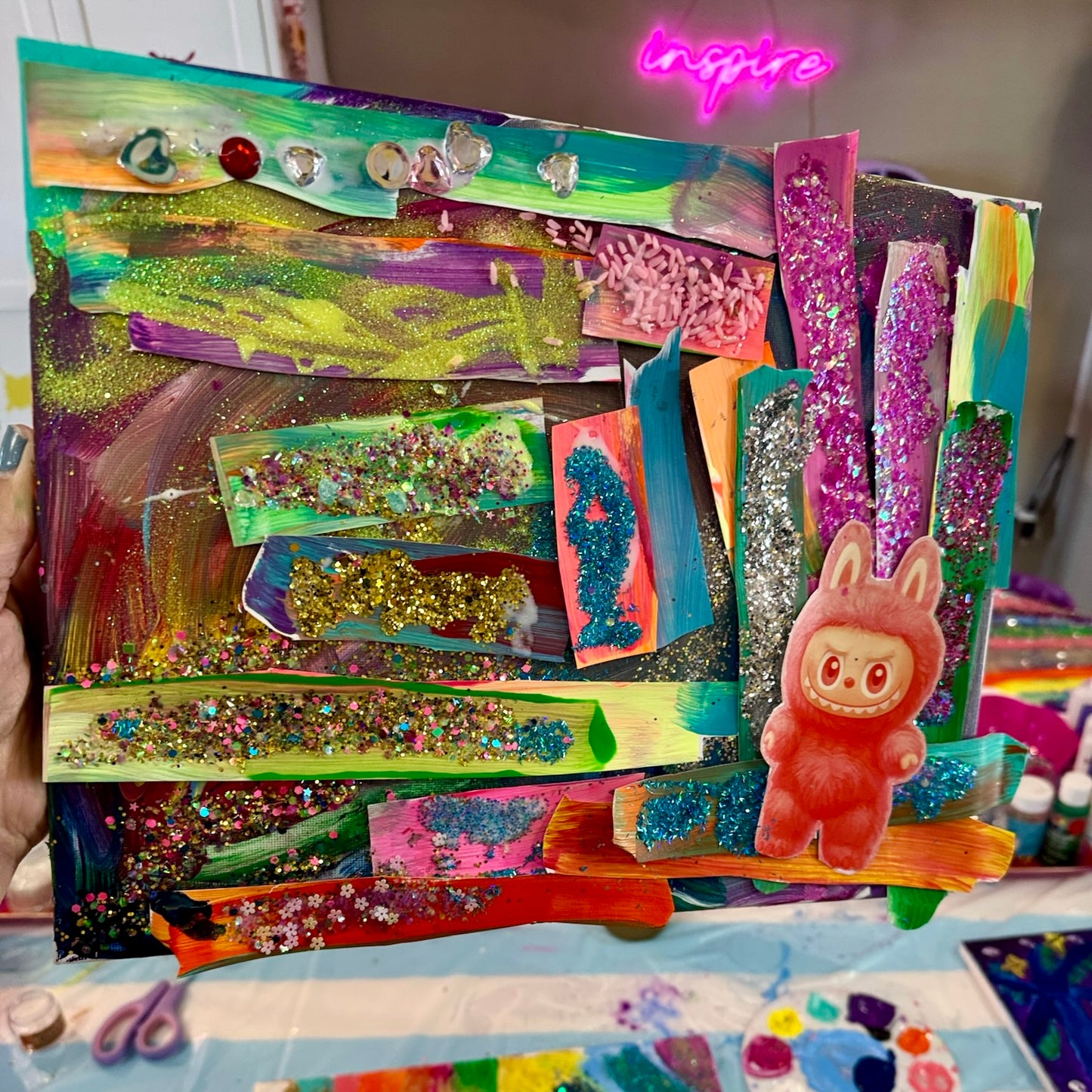 A colorful handmade abstract canvas art piece held by a hand, featuring layered glitter strips, paint splatters, rhinestones, and a small pink bunny figure. The background shows a craft table with paint palettes and supplies.