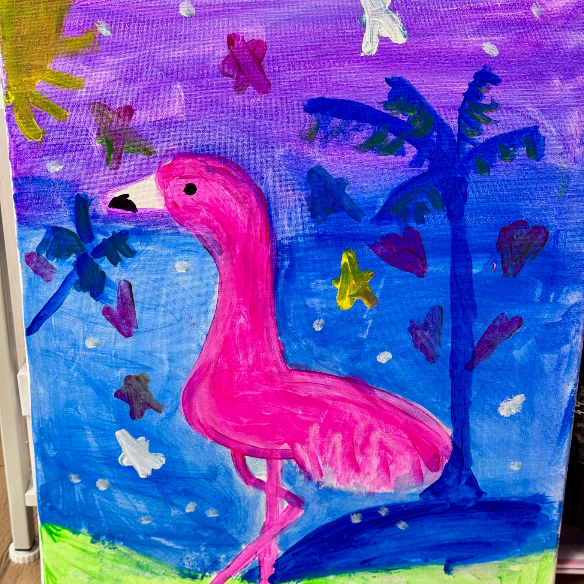 A colorful painted artwork featuring a bright pink flamingo standing on a blue and purple landscape with dark blue trees, green grass, and scattered white and yellow paint marks.