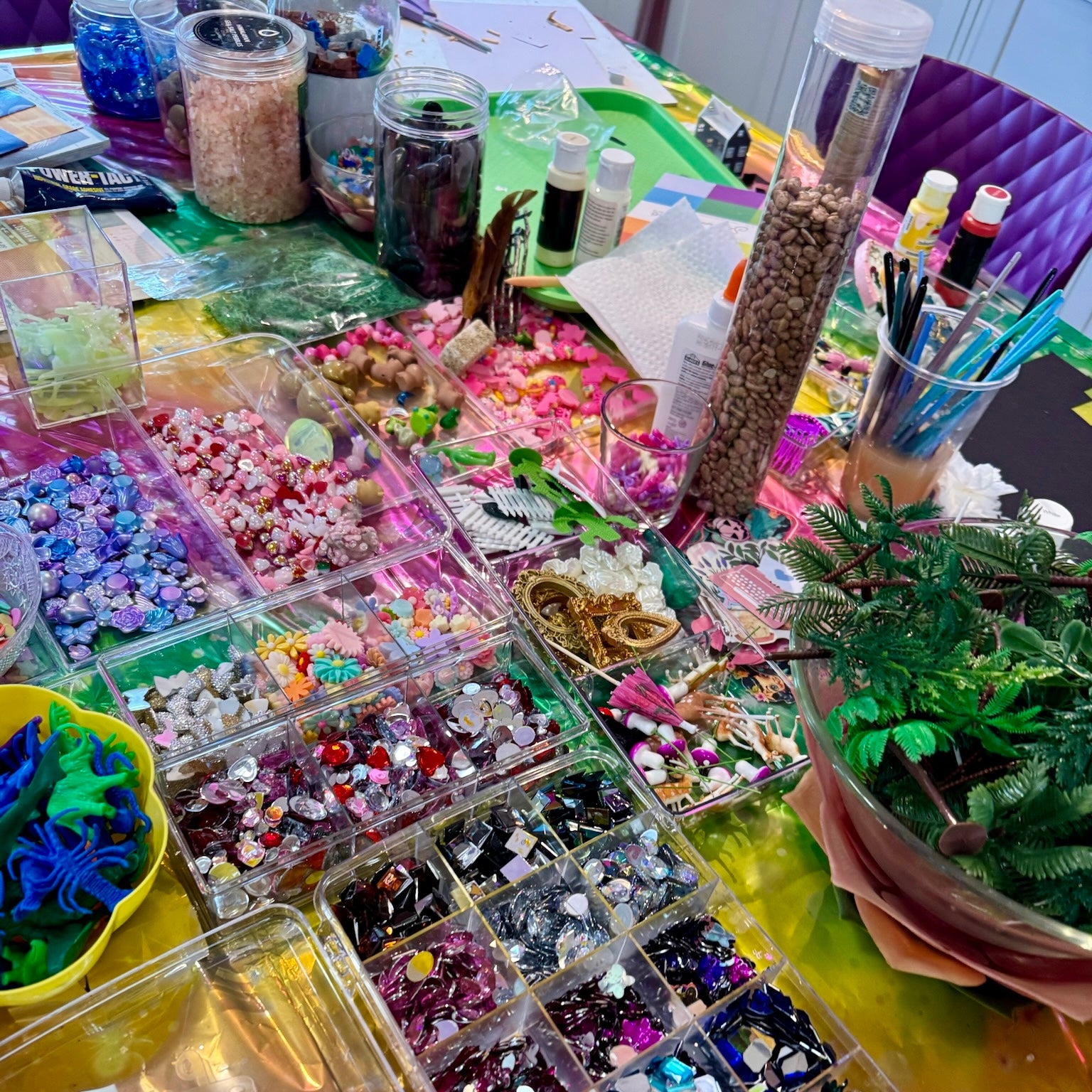 A tabletop display of many small clear organizer bins filled with colorful craft supplies, beads, rhinestones, glitter, paintbrushes, jars of loose materials, and a small potted green plant. No person or animal is visible, but this is not a clean single-product studio shot; it shows a styled craft supply arrangement in a real environment, so Lifestyle fits better than Product/Packaging.