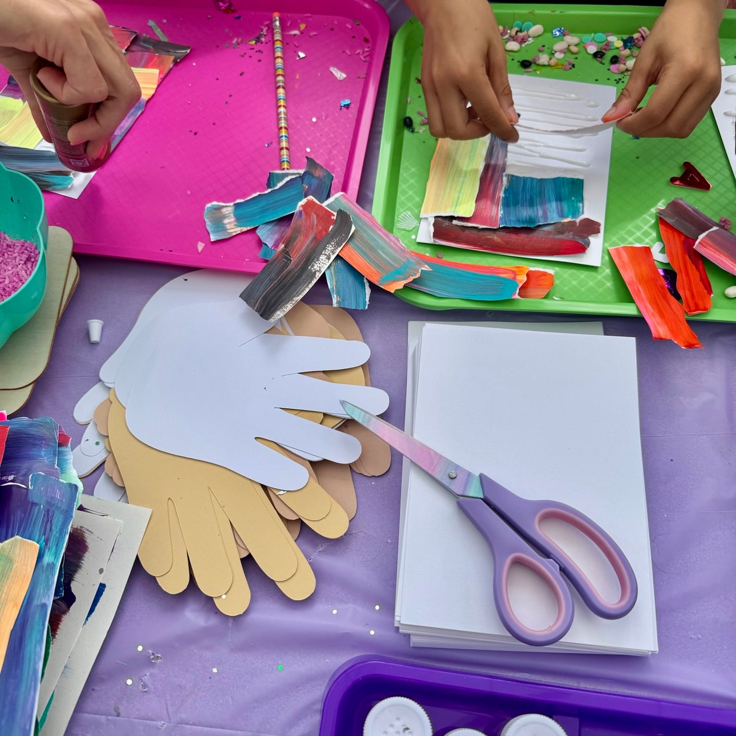 A craft activity setup on a purple tablecloth with hands visible, showing paper cutouts of hands in white and tan, colorful scrapbooking paper, scissors, glitter, beads, and small craft supplies.