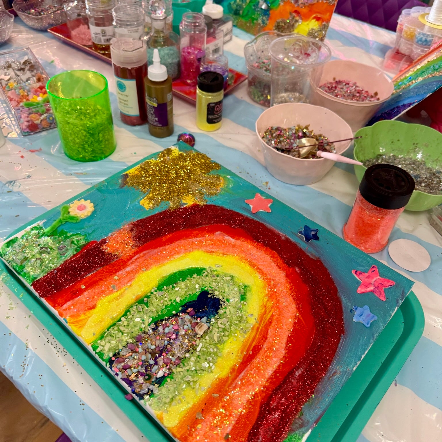 A colorful glitter rainbow art project on a teal tray, surrounded by craft supplies including bowls of glitter, paint bottles, and decorative stars on a craft table.