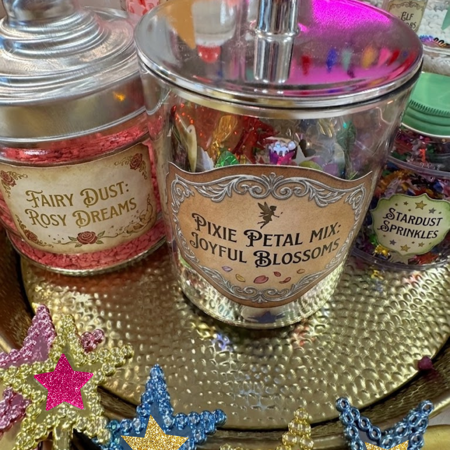 Decorative jars labeled 'Fairy Dust: Rosy Dreams' and 'Pixie Petal Mix: Joyful Blossoms' on a gold surface with star-shaped decorations.