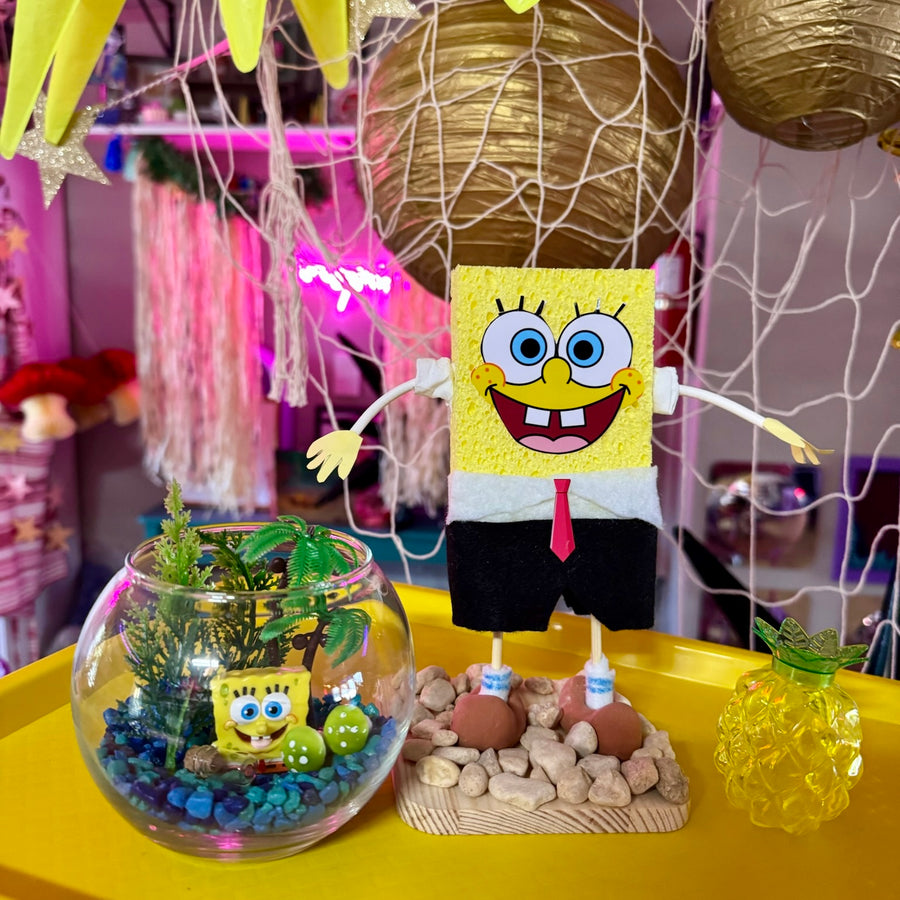 A handmade SpongeBob SquarePants plush toy made from textured yellow fabric, dressed in his white shirt and red tie, standing on a wooden base next to a glass terrarium with a mini SpongeBob figure and a yellow pineapple-shaped ornament.
